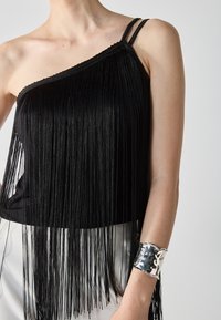 Svart ermeløs topp med asymmetrisk design og lange frynser; har tynne stropper og et strukturert sølvfarget mansjettarmbånd på håndleddet.