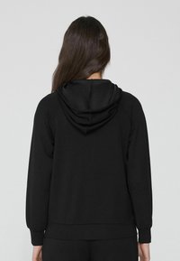 Černá mikina s kapucí, která má jemnou texturu, stahovací šňůrku a žebrované manžety. Navržena s volným střihem a minimalistikým stylem.