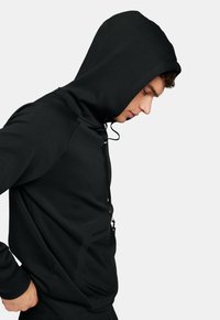 Hoodie noir avec un col montant et un cordon de serrage, doté d'une poche avant texturée et d'un tissu lisse. Conçu pour une silhouette ajustée.