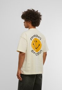 T-shirt en coton beige avec un grand graphique d'un visage souriant jaune et le texte noir "MARKET STUDIOS" dans le dos. Manches courtes, coupe décontractée.