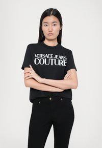 Juodas medvilninis marškinėliai su baltu "VERSACE JEANS COUTURE" logotipu ant priekio. Modelis nešioja juodas kelnes su aukso detalėmis.