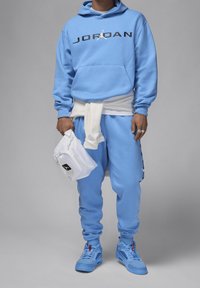 Sweat-shirt bleu clair avec logo "JORDAN" noir, pantalon de jogging assorti et baskets bleues. Sac blanc tenu à la main. Conception axée sur le confort.