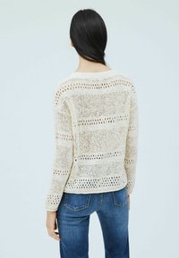 Ljust beige stickad tröja med ett öppet vävmönster och horisontella ränder, tillsammans med blå denimjeans.