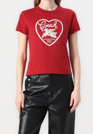 Vrouw draagt een rood T-shirt met een wit hart en een gevleugeld paard logo en een zwarte leren broek tegen een effen achtergrond.