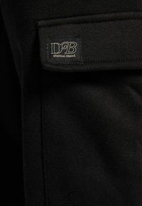 Svart tyg med en slät textur, med en rektangulär etikett i det övre vänstra hörnet som har "DB" och "eternal mood" i guldtext.