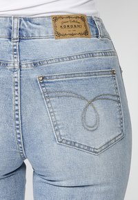 Jeans de mezclilla azul claro con una etiqueta marrón que dice "KOROSHI". Presentan un patrón de costura único en el bolsillo trasero y detalles de herrajes plateados.