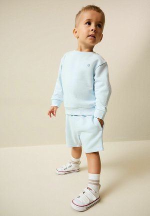 Mladý chlapec v svetlomodrej mikine a šortkách s bielymi ponožkami a teniskami na suchý zips, pozerajúci mierne nahor.