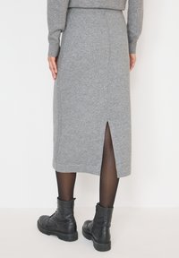 Femme debout portant une jupe midi en tricot grise avec fente au dos, des collants noirs transparents, des bottines en cuir noires et un pull gris assorti.