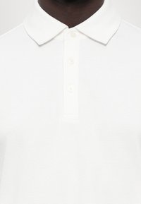 Hvit polo skjorte med et teksturert mønster, klassisk krage og tre knapper foran halsen, laget av lett stoff.