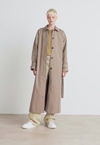 Lång beige trenchcoat med krage och svarta knappar. Bärs över en gul topp och krämfärgade byxor, tillsammans med vita sneakers.