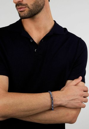 Bracciale di perline blu con tonalità variate di blu e una perla centrale in accent; indossato su un polso, in abbinamento a una polo nera.