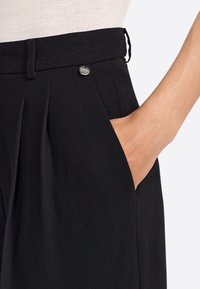Black tailored trousers with a pleated design, side pockets, and a silver button accent at the waistband. Smooth, textured fabric.