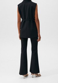 Schwarze ärmellose Tunika mit einem Gürtel in der Taille, kombiniert mit schwarzen ausgestellten Hosen. Das Outfit zeichnet sich durch eine glatte Textur und ein minimalistisches Design aus.