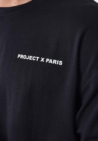 Zwarte katoenen t-shirt met witte tekst "PROJECT X PARIS" nabij de linkerschouder. Glad materiaal en klassieke pasvorm.