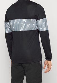 Černé atletické tričko s dlouhým rukávem, na zádech s vodorovným kovově stříbrným pruhem s abstraktními vzory. Hladká textura, běžný střih.