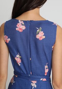 Blå blommig klänning med rosa blommor, ärmlös design, dragkedja i ryggen och ett knytbälte i midjan. Texturerad tyg.