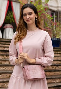 Rosa Crossbody-Tasche mit strukturiertem Krokodilmuster, goldenen Beschlägen und einem dekorativen Riemen. Das Modell trägt ein hellrosa Kleid mit Puffärmeln.