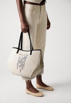 Lys beige kanvas tote bag med sort trim, som har en illustrasjon av en byscene og to figurer. Sorte håndtak for enkel bæring.