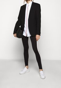 Blazer nero su una camicia bianca con scritte rosse, abbinato a leggings neri con un dettaglio a righe bianche e sneakers bianche.