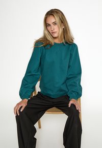 Felpa teal a girocollo con maniche a sbuffo e polsini a costine, abbinata a pantaloni neri a gamba larga, modello seduto su una sedia.