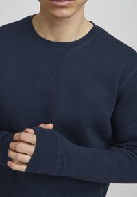 Jūras spēku garenrīkota kreklai ar teksturētu audumu, ribotu kakla izgriezumu un pārklājošiem šuvju elementiem. Arī aproču daļā ir iegriezumi pirkstiem.