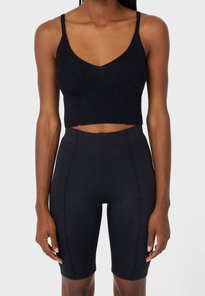 Sort slim fit ribbet crop-top med tynde stropper kombineret med højtaljede sorte cykelshorts på en stående person.