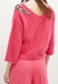 Vaaleanpunainen V-kaula-aukkoinen neulepehmeästä, pörröisestä materiaalista, jossa vasemmalla olkapäällä koristeellisia jalokiviyksityiskohtia ja resorireuna.