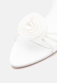 Άσπρο παπούτσι με μυτερή μύτη, διακοσμημένο με λεπτομέρεια τριαντάφυλλου και λεπτές λουράκια σε ανοιχτό φόντο.