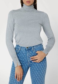 Maglione a collo alto grigio chiaro con texture a coste, maniche lunghe con dettagli di bottoni, abbinato a jeans blu a vita alta con dettagli a fantasia.