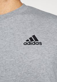 Šedé bavlnené tričko s čiernym vyšitým logom Adidas a tromi diagonálnymi pruhmi. S výstrihom do okrúhla, jemná textúra.