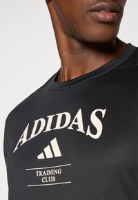 Black athletic t-shirt featuring "ADIDAS" logo in cream, with "TRAINING CLUB" text, made from a smooth, breathable material. Short sleeves.