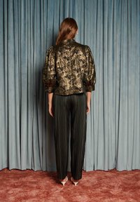 Woman stands facing blue curtain wearing gold floral textured puff-sleeve jacket, black pinstripe trousers, and white pointed heels.