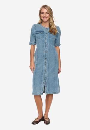 Kvinde smilende, iført en midt-lang denimkjole med knapper og albuelange ærmer samt sorte vævede flade sko, stående mod en hvid baggrund.