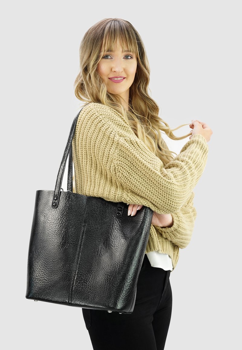 Zwarte leren totebag met textuur en twee korte handvatten, met een gestructureerd ontwerp en drukknoopaccenten, vastgehouden door een persoon in een beige trui.