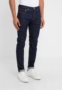 Muž ve tmavě modrých úzkých džínách vyhrnutých u kotníků, s bílými teniskami a bílými ponožkami, stojící před jednobarevným pozadím.