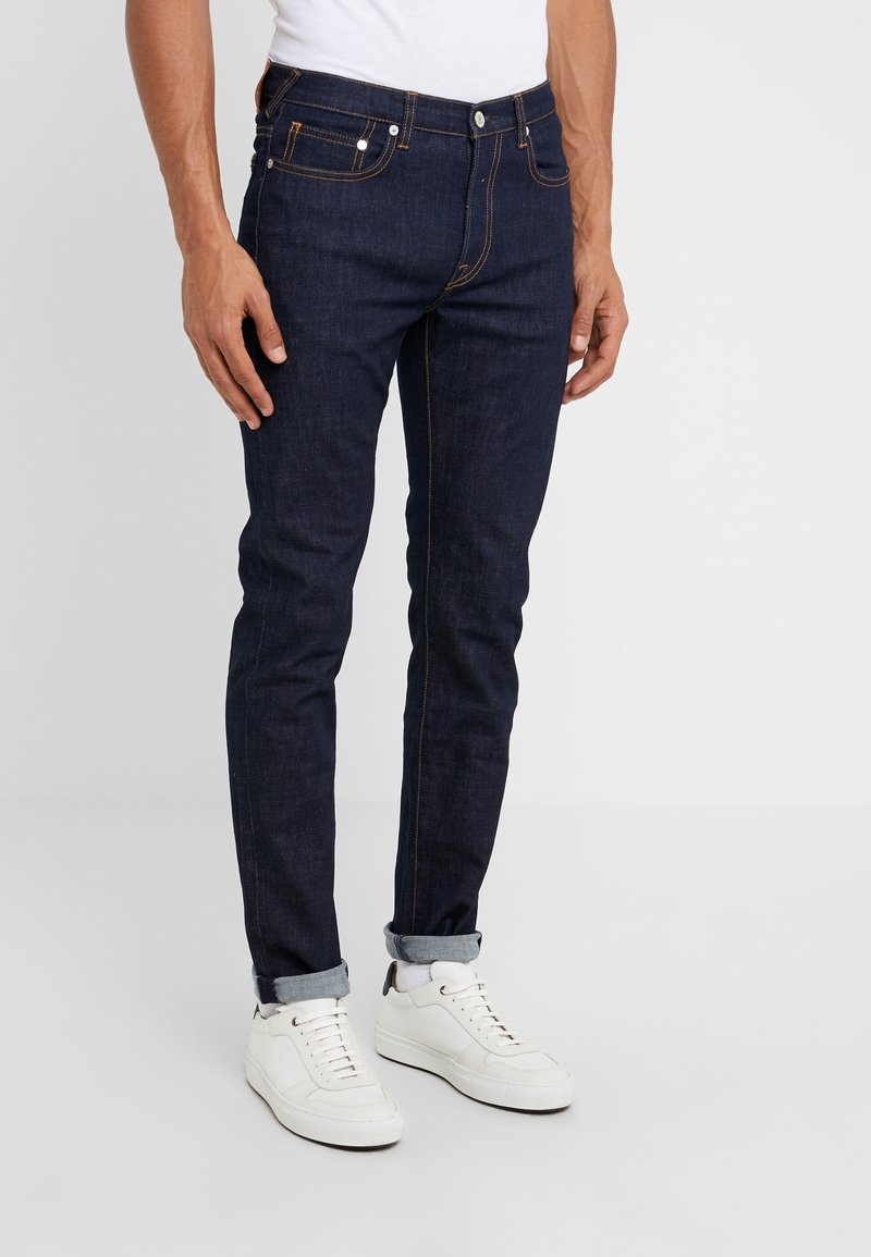 Muž ve tmavě modrých úzkých džínách vyhrnutých u kotníků, s bílými teniskami a bílými ponožkami, stojící před jednobarevným pozadím.