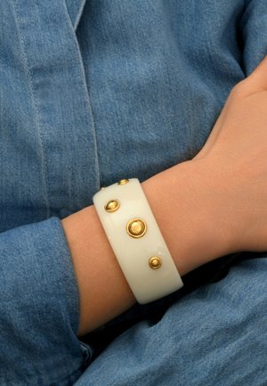 Bracelet à bangle blanc avec des accents dorés circulaires, surface lisse et brillante, porté sur un poignet contre un tissu en denim.