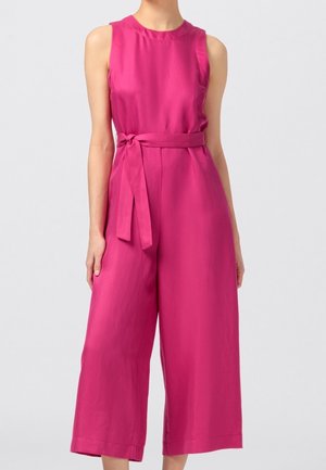 Femme portant une combinaison sans manches rose vif à jambes larges avec une ceinture nouée à la taille, debout devant un fond clair uni.