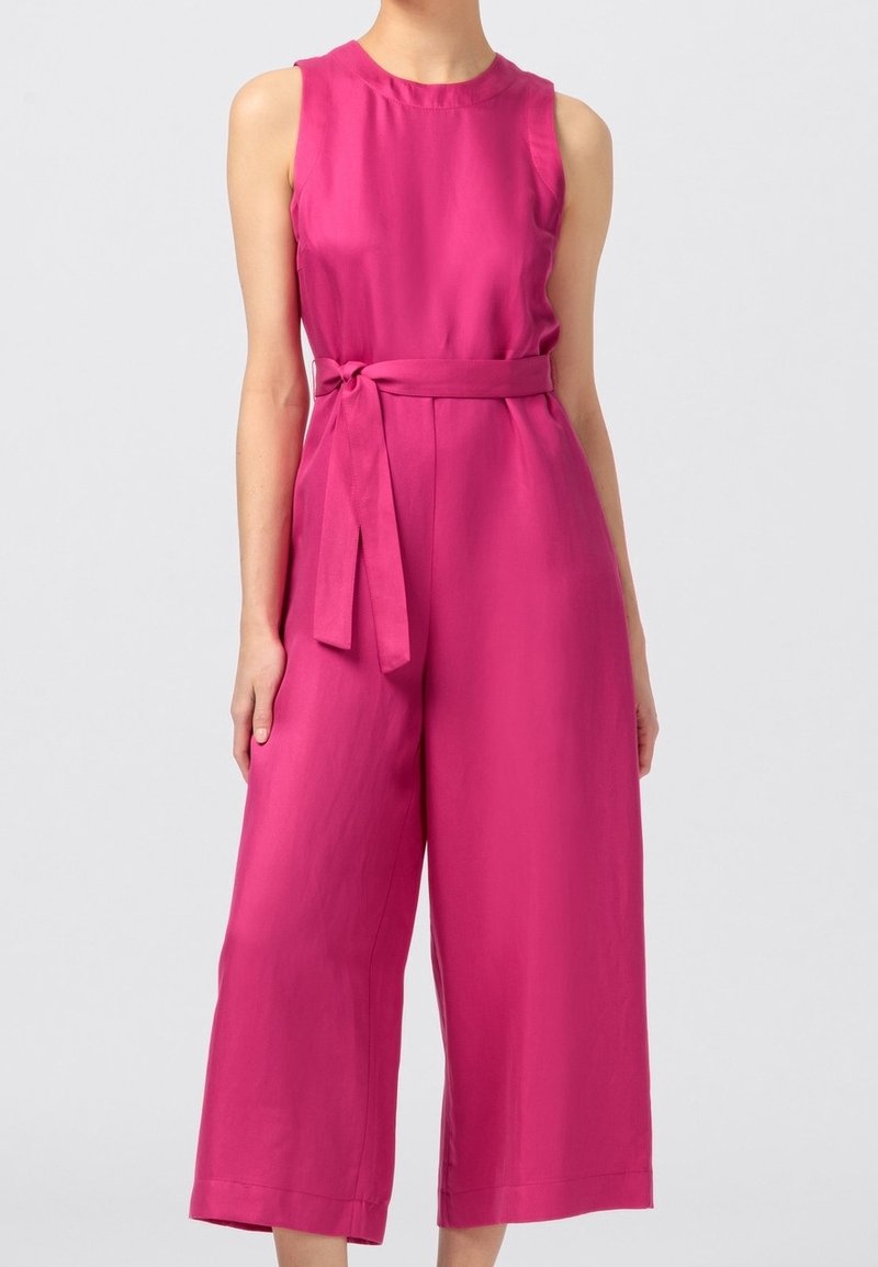 Femme portant une combinaison sans manches rose vif à jambes larges avec une ceinture nouée à la taille, debout devant un fond clair uni.