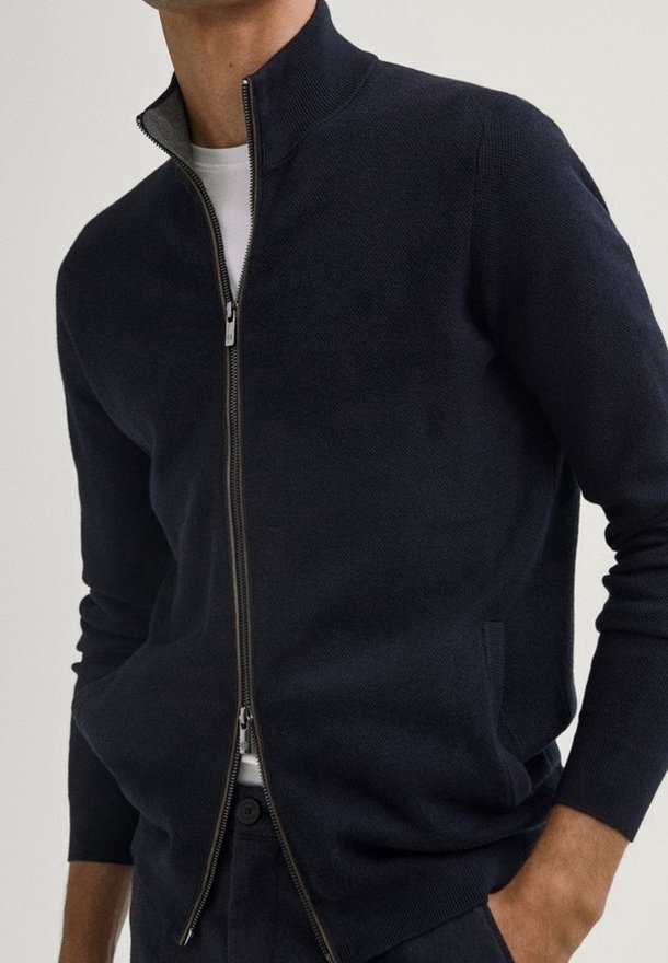 Homme portant un pull zippé bleu marine foncé par-dessus un t-shirt blanc, les mains dans les poches, visible du cou jusqu’aux hanches.
