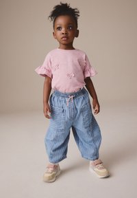 Camisetta rosa a maniche corte con bordi arricciati e dettagli ricamati, abbinata a pantaloni cargo a righe blu larghi e sneaker multicolori.