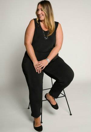 Combinaison noire avec un design sans manches, en tissu lisse et coupe décontractée. Accentuée par un collier circulaire et portée avec des ballerines noires.