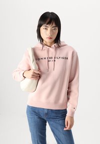 Hoodie en coton rose avec texte logo, poignets et ourlet côtelés. Associé à un jean bleu et tenant un sac à bandoulière blanc.