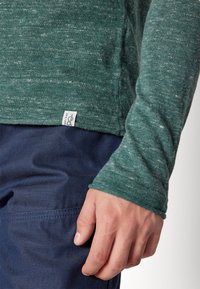 Grön texturerad långärmad skjorta med rundad nederkant, ribbade ärmslut och en liten etikett i nederkanten. Bärs med mörkblå byxor.