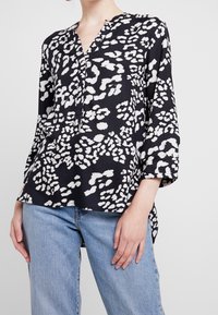 Czarna bluzka z białym abstrakcyjnym wzorem, dekolt w serek, rękawy 3/4 i luźny krój, w połączeniu z jasnoniebieskimi dżinsami.