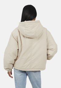 Beżowa pikowana kurtka z przestronnym kapturem, elastycznymi mankietami i luźnym krojem. Materiał wydaje się miękki w dotyku i ma gładką fakturę. Pokazano widok z tyłu.