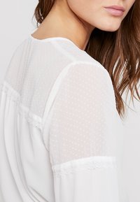 Blusa bianca con maniche trasparenti a pois, rifiniture in pizzo e scollatura rotonda. Il tessuto ha una consistenza liscia con un aspetto leggero e arioso.