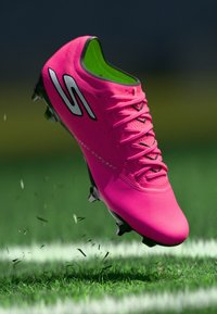 Pinkfarbener Fußballschuh mit einer strukturierten Oberfläche, schwarzen Schnürsenkeln und grünem Innenfutter. Verfügt über ein Logo und Stollen, die für Traktion auf Gras ausgelegt sind.