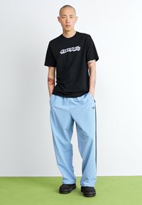 Camiseta preta com gráfica branca, calças atléticas azuis com riscas laterais e sapatos pretos, exibidos contra um fundo liso.