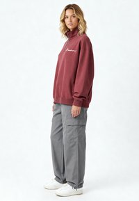 Sweat-shirt bordeaux à demi-fermeture éclair avec des détails en script blanc, associé à un pantalon cargo gris clair et des baskets blanches. Tissu doux, coupe décontractée.